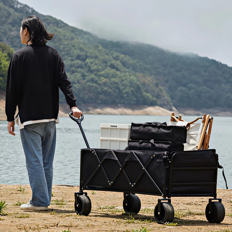 Cargue la imagen en el visor de la galería, Carrito vagón plegable multiusos FilmCrew. Persona jalando un carrito vagón plegable negro FilmCrew cargado con equipo de camping a la orilla de un lago.
