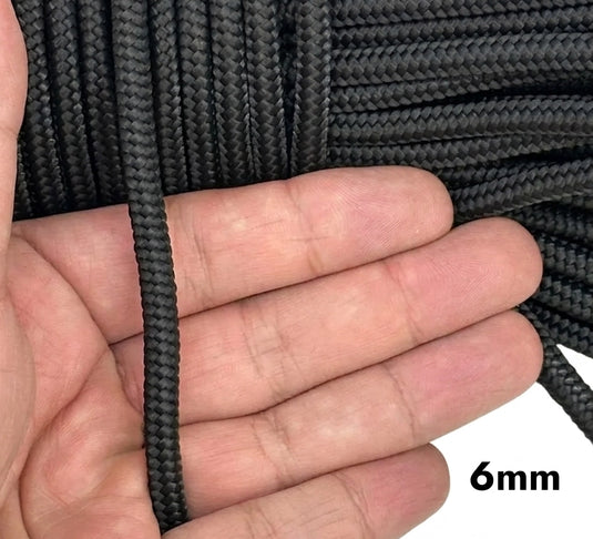 Piola cuerda de polipropileno negra.  Mano sosteniendo un extremo de cuerda negra de 6 milímetros de grosor junto a madeja para mostrar su grosor, textura y proporción.